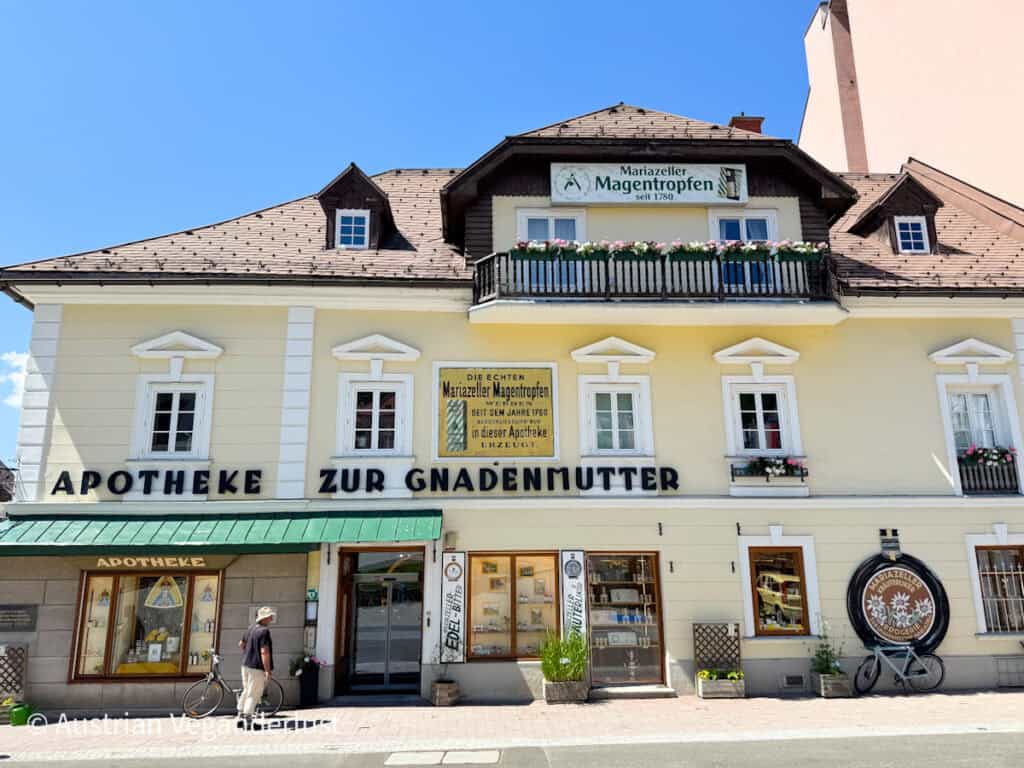 Die historische "Apotheke zur Gnadenmutter" in Mariazell mit gelber Fassade, Blumenkästen auf dem Balkon und Schildern, die für das traditionelle Kräuterheilmittel "Mariazeller Magentropfen" werben.