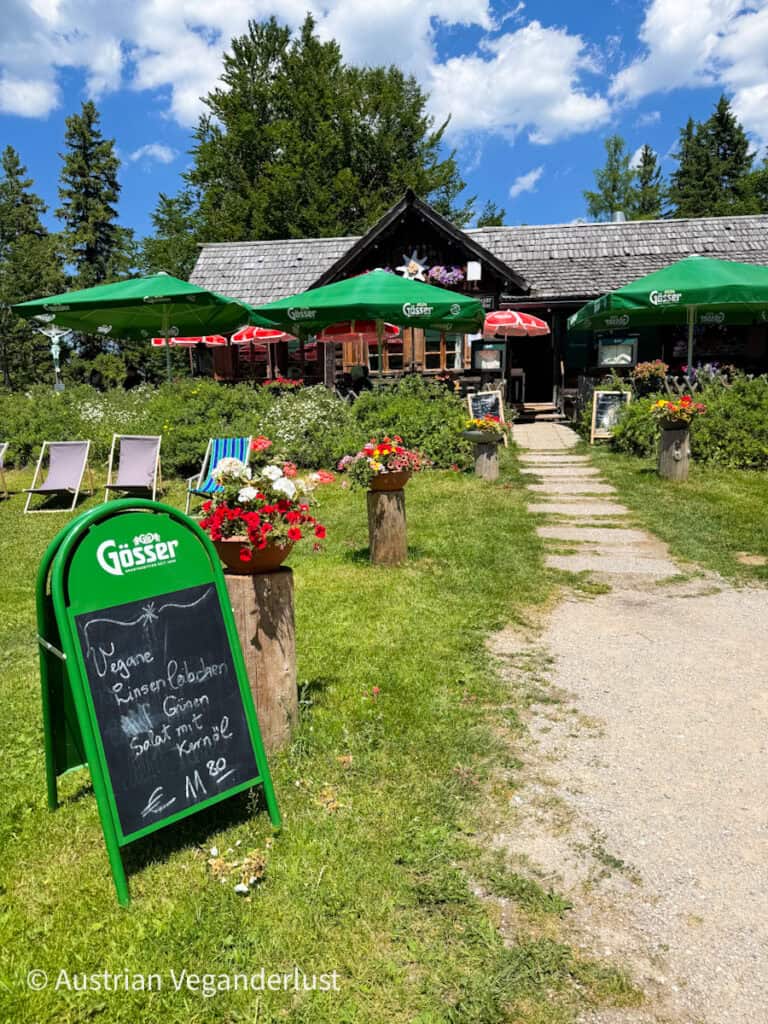 Ein gemütliches Hüttenrestaurant mit grünen Gössser-Schirmen und roten Blumentöpfen draußen, mit einem Kreideschild, auf dem steht: "Vegane Linsenlaibchen, grüner Salat mit Kernöl €11,80".
