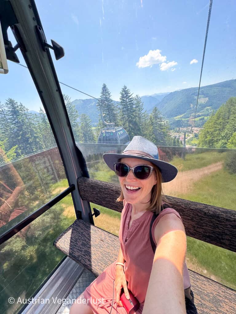 Eine Frau in einem ärmellosen Kleid und einem Sonnenhut lächelt in einer Gondelbahn, während sie einen Panoramablick auf bewaldete Berge und einen klaren Sommerhimmel hinter sich hat.