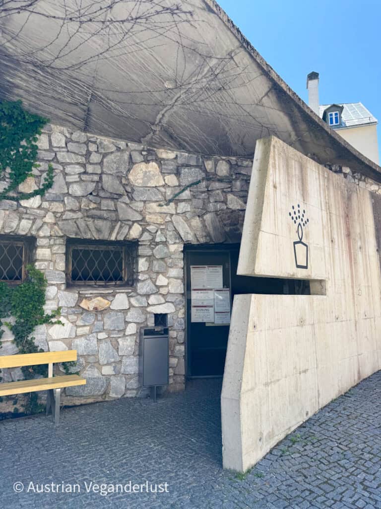 Außenansicht des Kerzenheiligtums in Mariazell aus Stein und Beton mit einem kantigen Dach und mit Wein bewachsenen Wänden, neben einer gelben Bank und Informationsschildern.