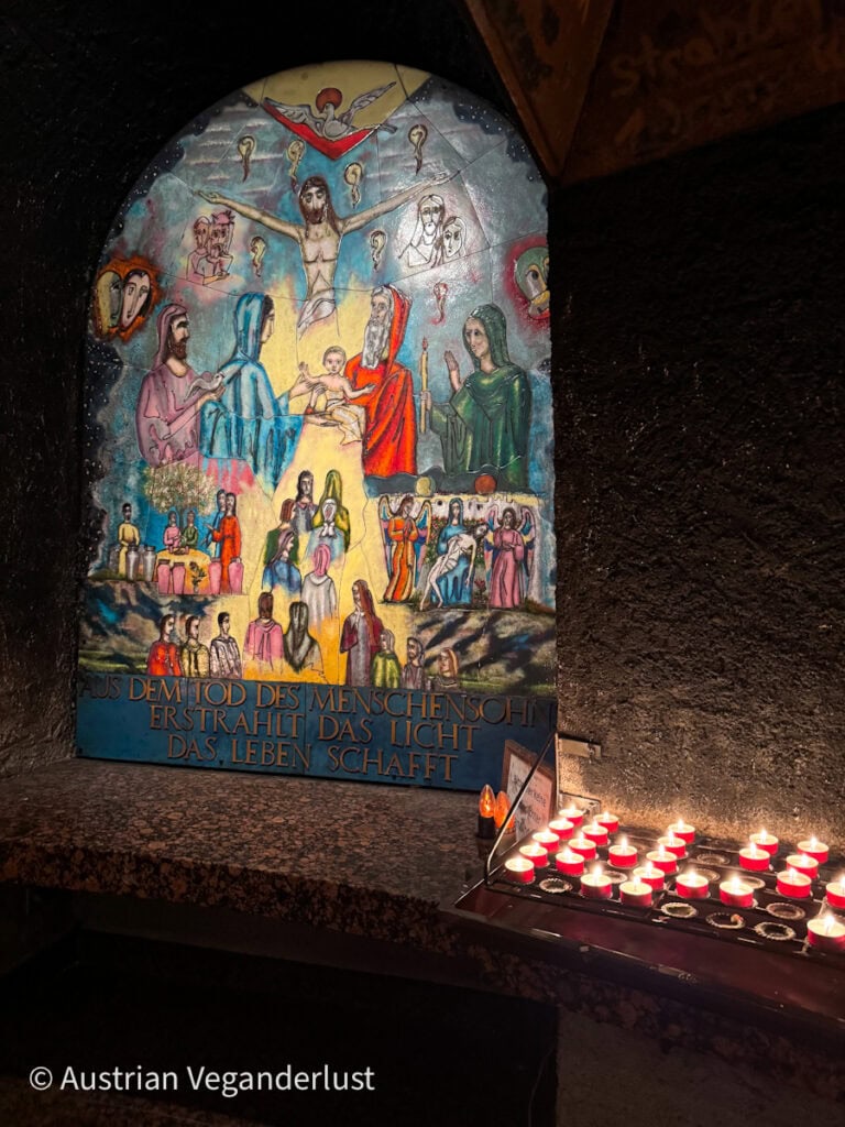 Ein farbenfrohes religiöses Wandgemälde, das Jesus und verschiedene Heilige zeigt, leuchtet im warmen Licht neben einem Tablett mit roten Votivkerzen in einer schummrigen Kapelle.