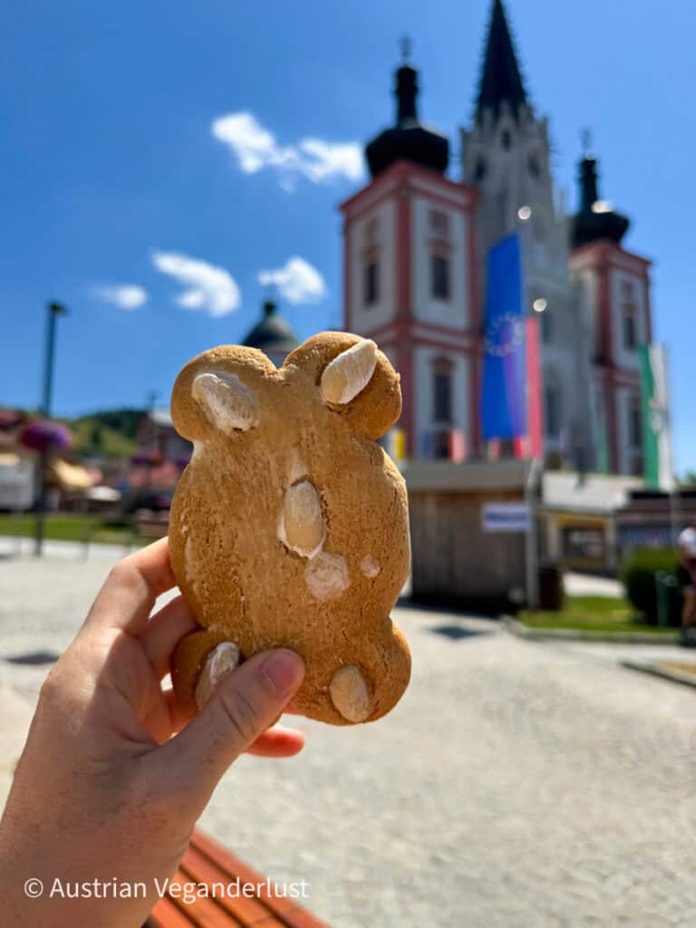 Eine Hand hält einen Lebkuchen in Bärenform mit Mandelakzenten hoch, während die Mariazeller Basilika im sonnigen Hintergrund verschwommen zu sehen ist.