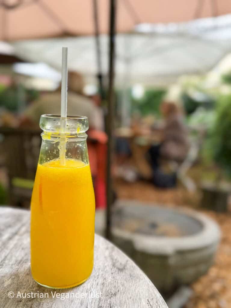 Eine Glasflasche, gefüllt mit leuchtend gelbem Saft und einem Strohhalm, steht auf einem verwitterten Holztisch in einem Gartencafé mit weichgezeichneten Gästen im Hintergrund.