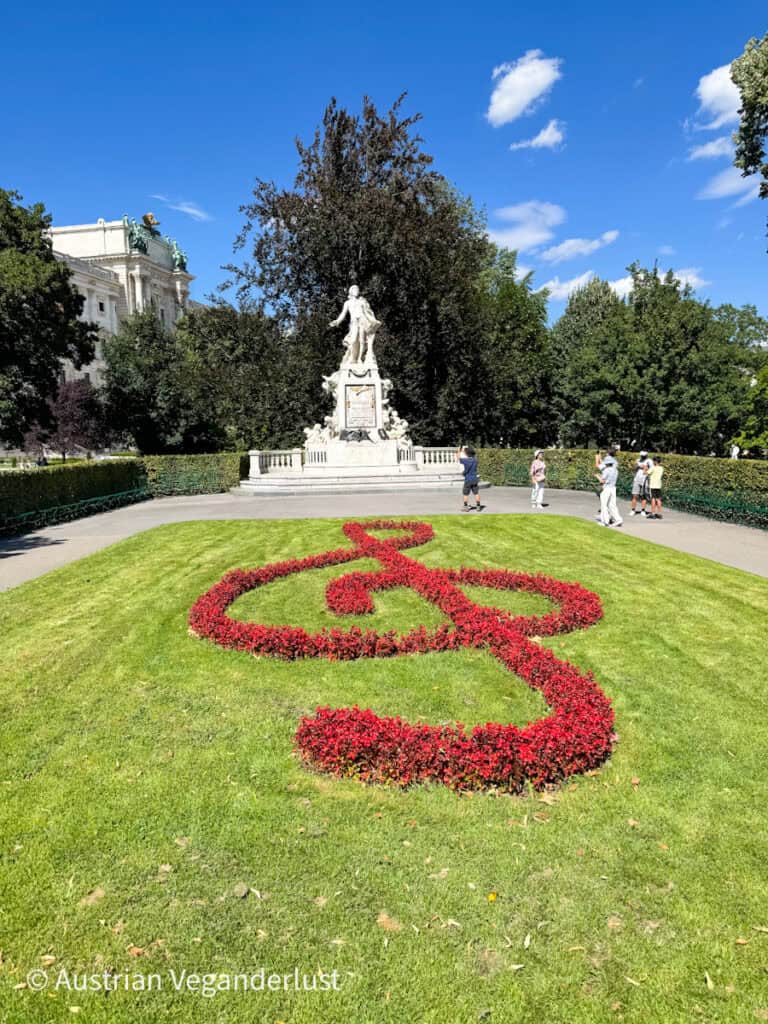Eine weiße Marmorstatue von Mozart steht in der Mitte hinter einem Beet aus roten Blumen, das die Form eines Violinschlüssels hat, auf einer grünen Wiese im Burggarten in Wien. Man sieht Touristen, die unter einem strahlend blauen Himmel Fotos machen.