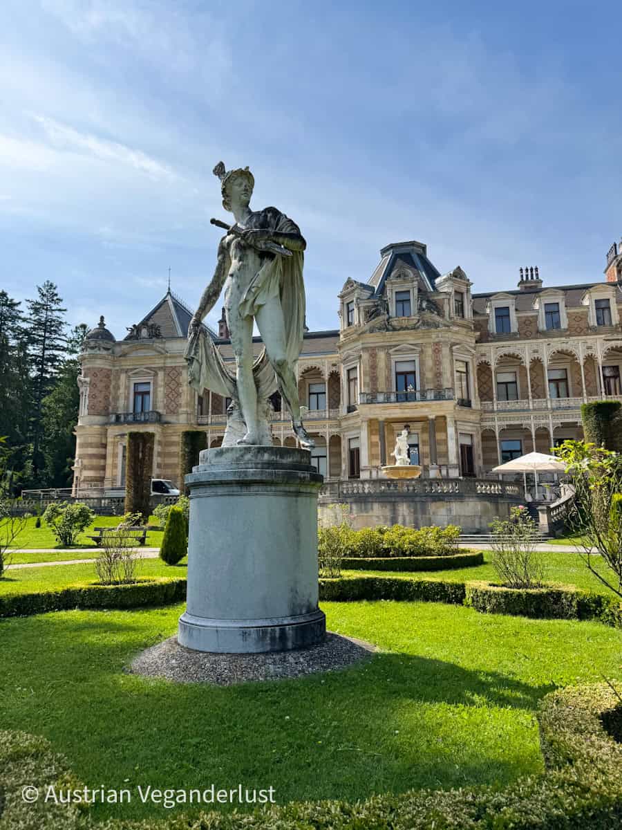 Marble statue of a classical figure stands on a pedestal in the manicured gardens of the Hermesvilla, a historic palace in Vienna. The elegant villa, with its turrets and balconies, serves as a regal backdrop under a clear blue sky.