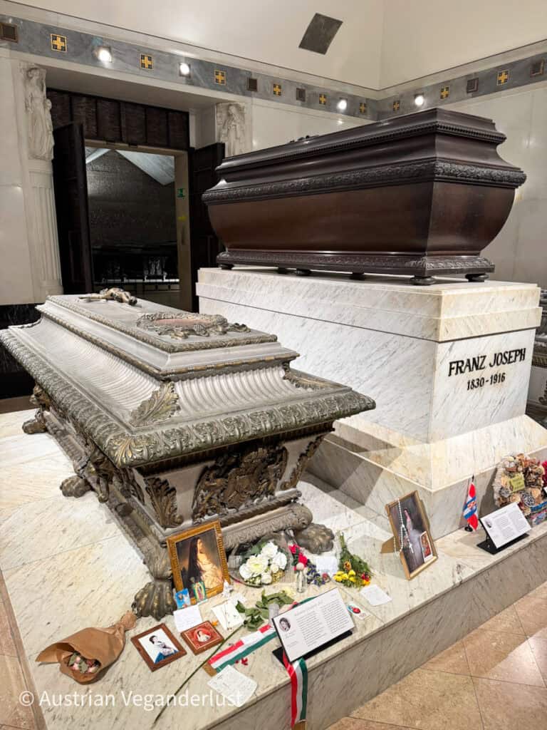 Elaborate imperial sarcophagi of Emperor Franz Joseph and Empress Elisabeth displayed side by side in the Capuchin Crypt in Vienna. The tombs rest on marble platforms adorned with flowers, portraits, and tribute notes left by visitors.