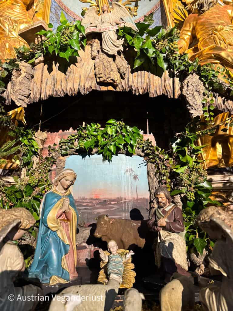 A traditional Nativity scene with figurines of Mary, Joseph, and baby Jesus lying in a manger, surrounded by animals and greenery. Above the stable, an angel holds a banner reading “Gloria in Excelsis Deo,” framed by golden details and ivy decorations.