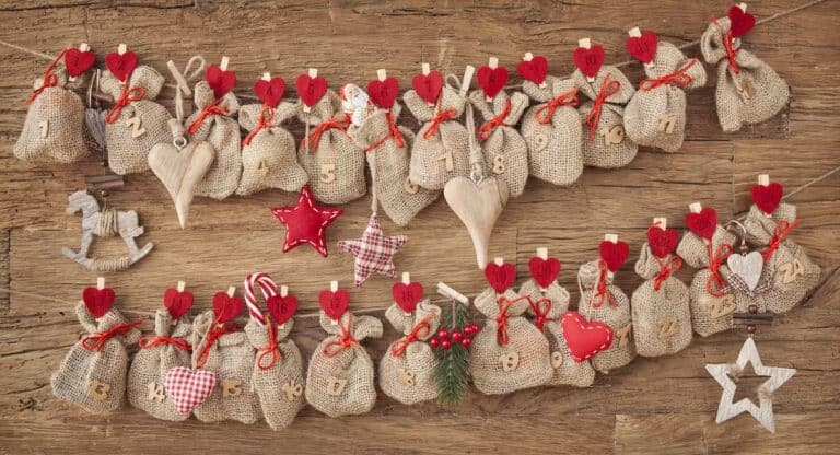 A rustic homemade Advent calendar made of small burlap bags numbered 1 to 24, each tied with red string and clipped with red felt hearts. The bags are strung on twine against a wooden background, decorated with small stars, hearts, and festive ornaments.