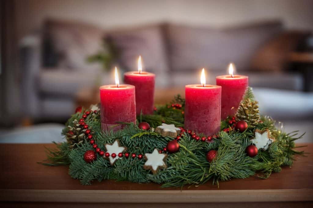 An Advent wreath decorated with pine branches, red beads, pinecones, and star-shaped cookies, holding four red candles, each lit to create a warm festive glow. The wreath sits on a wooden table with a blurred sofa in the background.