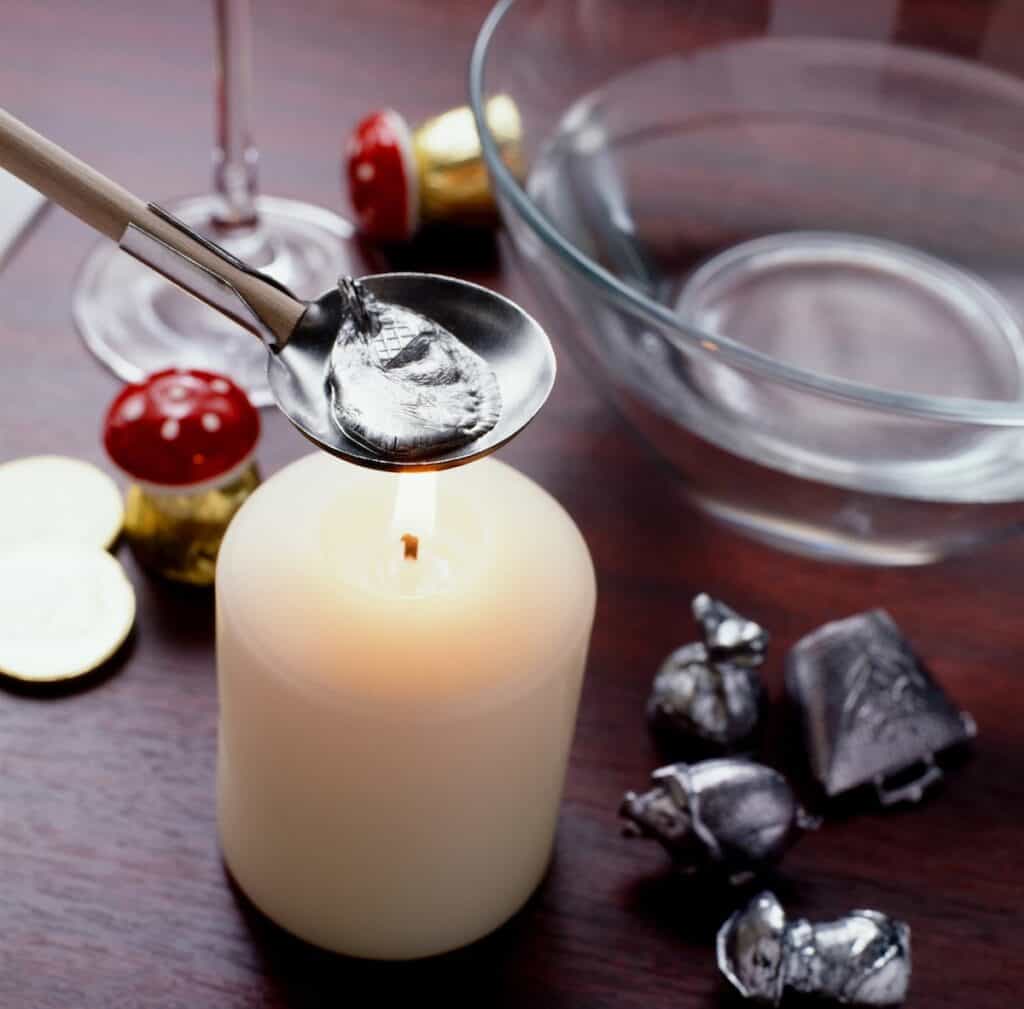 A close-up of the traditional Austrian New Year’s custom of Bleigießen, where molten lead or tin is melted in a spoon over a candle flame before being dropped into cold water. Small figurines and a bowl are nearby, ready for fortune-telling interpretations.