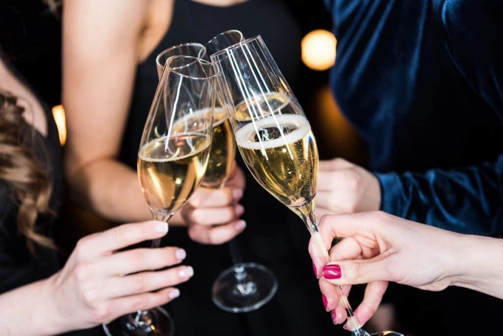 A close-up of people clinking champagne glasses in a festive toast, with golden sparkling wine bubbling in tall flutes. The elegant setting and evening attire suggest a New Year’s or celebratory occasion.