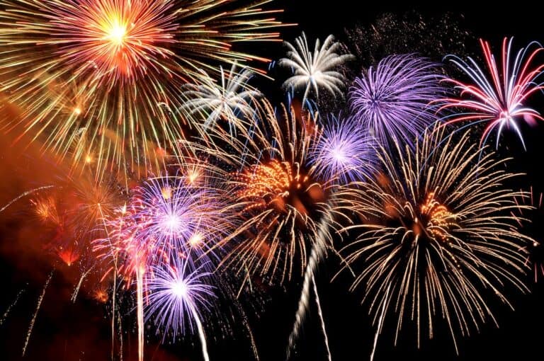 A night sky filled with vibrant fireworks bursting in red, gold, purple, and white, creating a dazzling display of light and color. The overlapping patterns capture the excitement of a festive celebration like New Year’s Eve.
