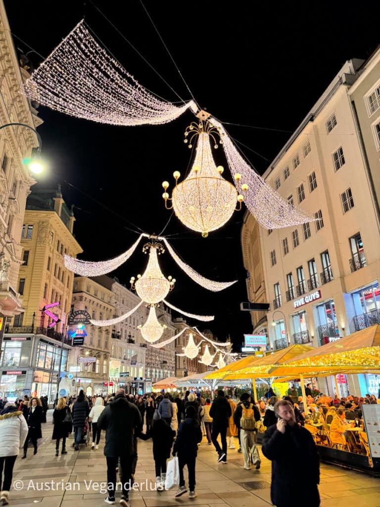 Vienna’s Graben street at night, glowing with cascading chandelier-style Christmas lights overhead and lined with busy market stalls under yellow-lit canopies. Shoppers and diners fill the bustling pedestrian zone, creating a vibrant holiday scene.