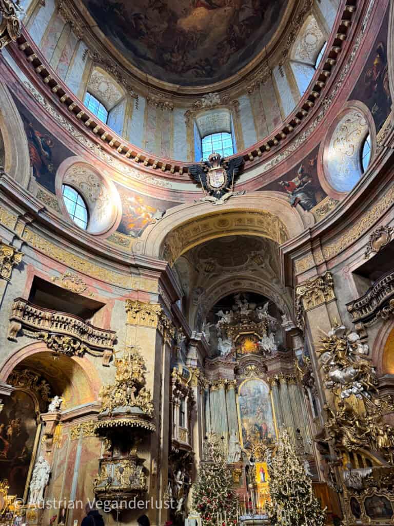 Interior of St. Peter’s Church in Vienna, richly decorated with gilded baroque sculptures, marble columns, and intricate frescoes on the dome above. The altar is surrounded by Christmas trees and glowing lights, adding festive warmth to the grandeur of the sacred space.