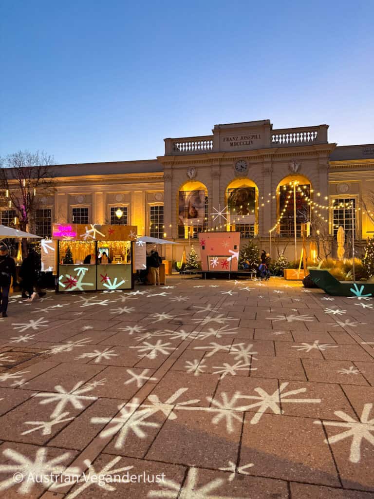 Die Weihnachtsmarktstände vor dem Wiener MuseumsQuartier erstrahlen bei Einbruch der Dunkelheit in goldenem Licht. Dekorative sternförmige Lichter werden auf den Bürgersteig projiziert und verleihen der eleganten Architektur der Franz-Joseph-I-Fassade eine verspielte Weihnachtsatmosphäre.