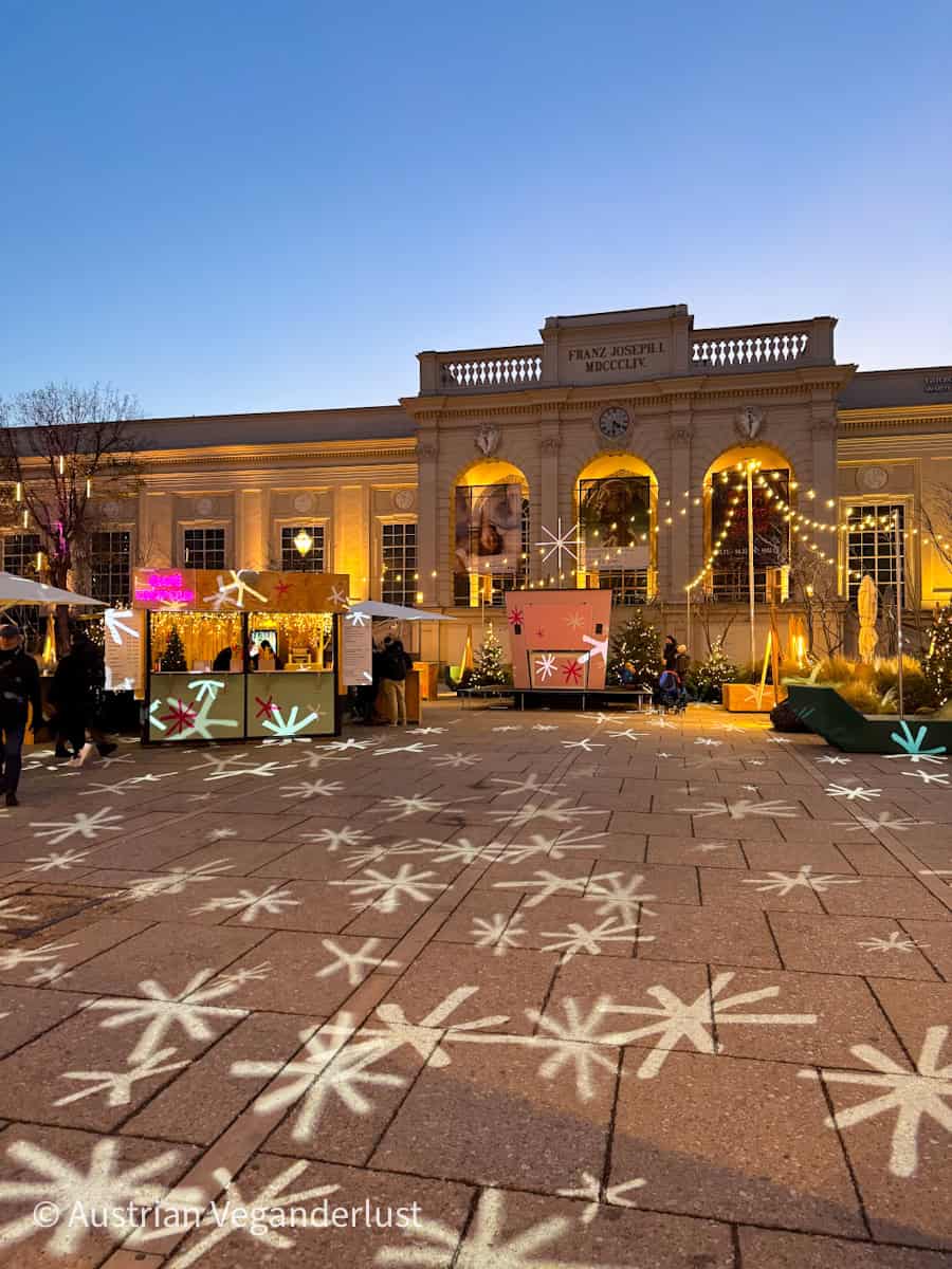 Christmas market stalls set up in front of Vienna’s MuseumsQuartier, glowing under golden lights as dusk settles in. Decorative star-shaped lights are projected onto the pavement, adding a playful holiday atmosphere to the elegant architecture of the Franz Joseph I façade.