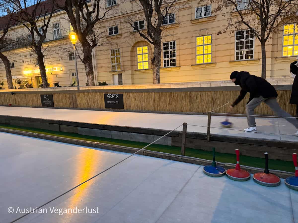 A person playing Eisstockschießen, or Bavarian curling, on an outdoor ice lane in Vienna at dusk, with colorful curling stocks lined up in the foreground. Behind the player, a historic cream-colored building is warmly lit, and bare trees line the edge of the rink.