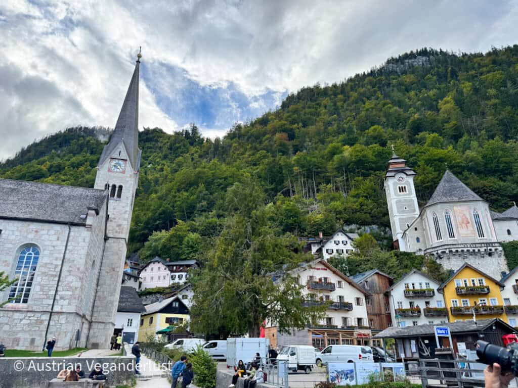Blick auf die Gebäude des Dorfes Hallstatt mit ihren bunten Fassaden und dem hohen Turm der evangelischen Kirche, umgeben von dicht bewaldeten Hügeln.