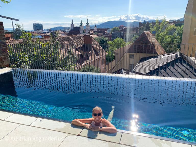 Eine Frau genießt einen Infinity-Pool auf dem Dach mit einem atemberaubenden Panoramablick auf das Grazer Stadtbild, die Kirchtürme und die fernen Hügel unter einem strahlenden Sonnenhimmel.