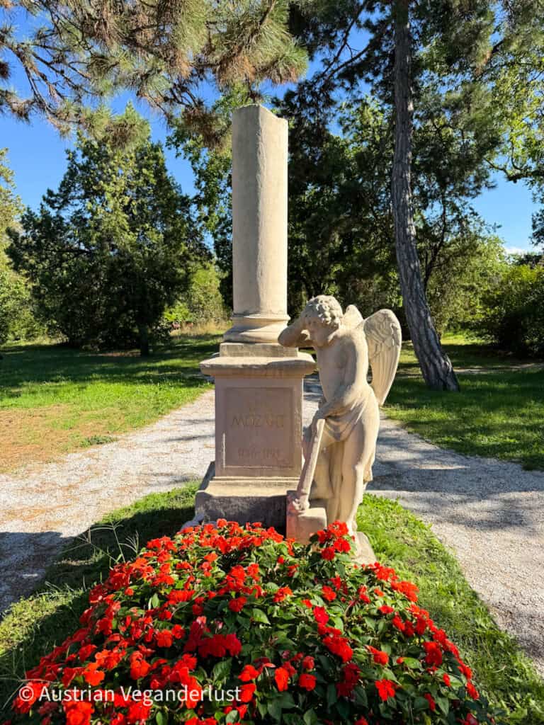 A stone monument dedicated to W.A. Mozart stands in a cemetery garden, featuring a broken column and a weeping angel statue surrounded by bright red flowers.