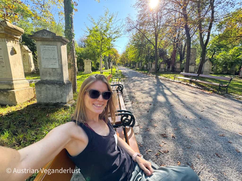 A smiling woman wearing sunglasses takes a selfie while seated on a bench along a sunlit cemetery path lined with gravestones and autumn trees.