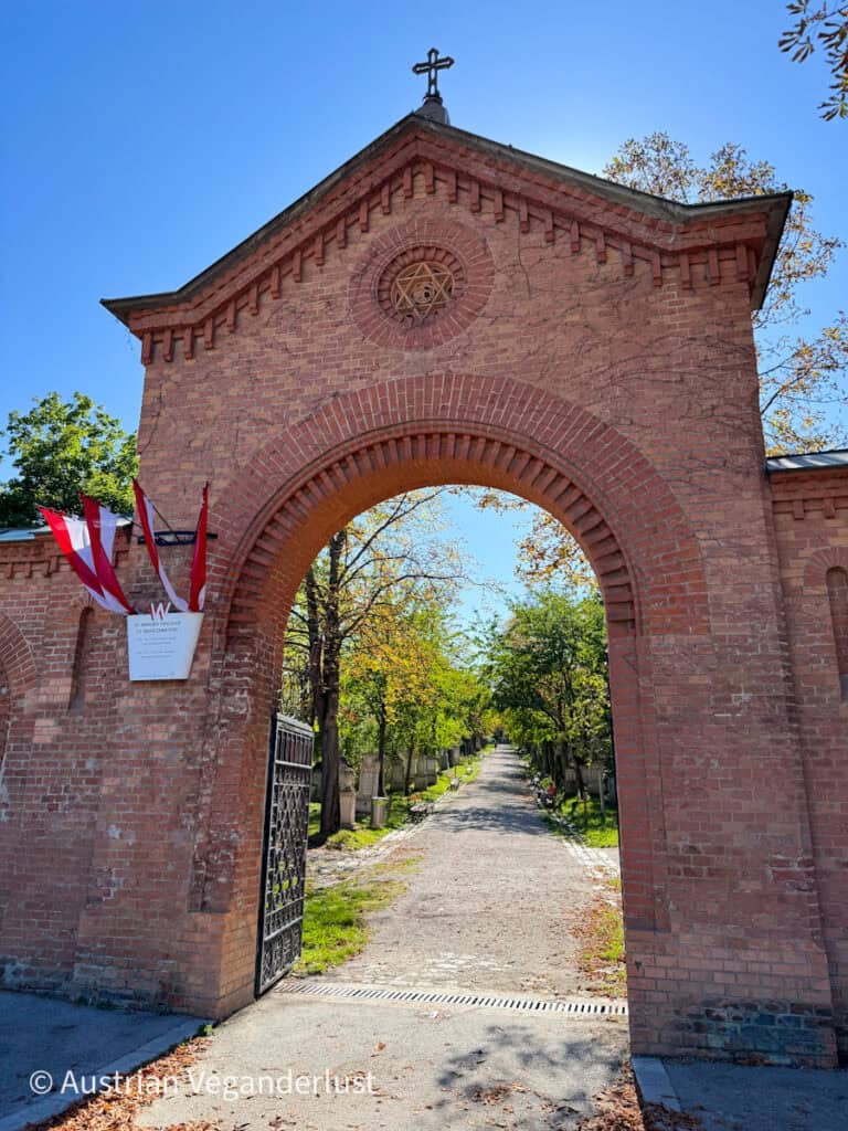 A large red-brick archway with a cross on top marks the entrance to Vienna’s Central Cemetery, leading to a long tree-lined path of gravestones.