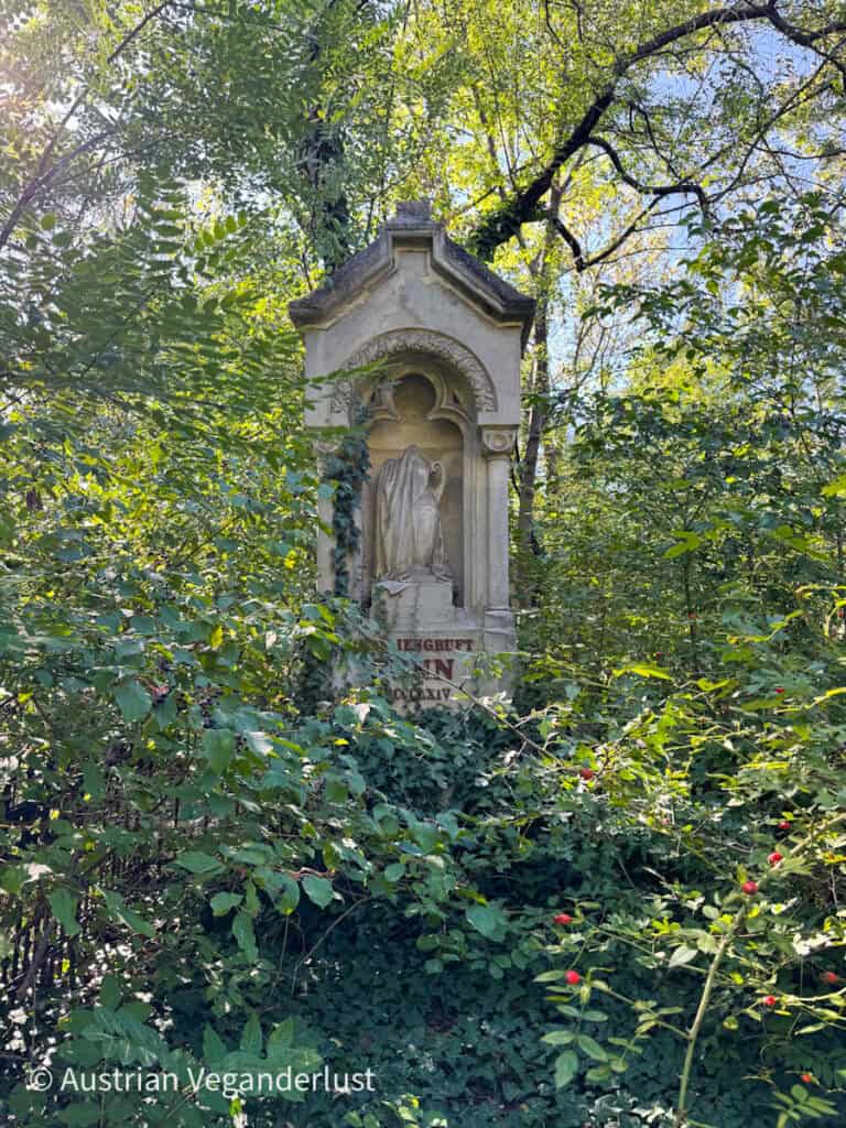 Ein steinernes Grabdenkmal, das teilweise von dichtem Grün und Efeu verdeckt ist, zeigt eine gewandete Figur im Relief, die sich in die natürliche Umgebung einfügt.