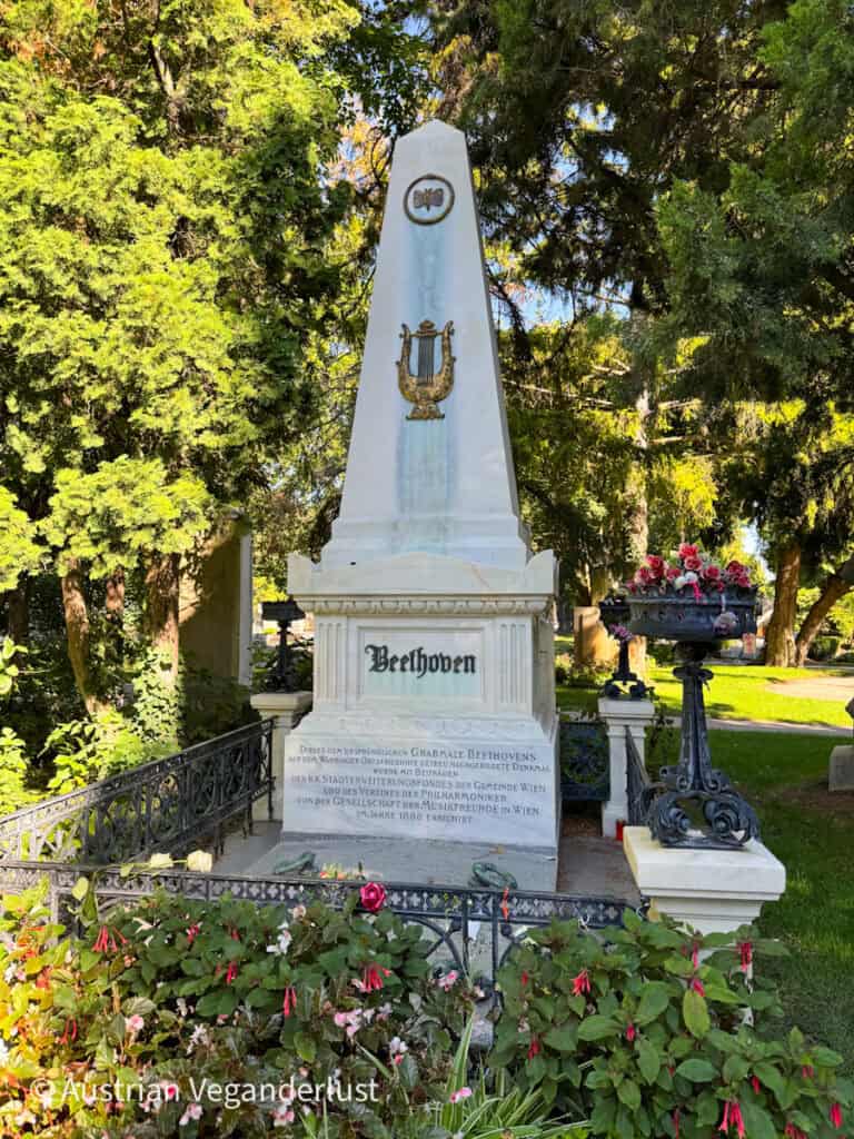 Weißer Obelisk-Grabstein des Komponisten Ludwig van Beethoven mit einem goldenen Leier-Emblem, umgeben von Blumen und Bäumen.