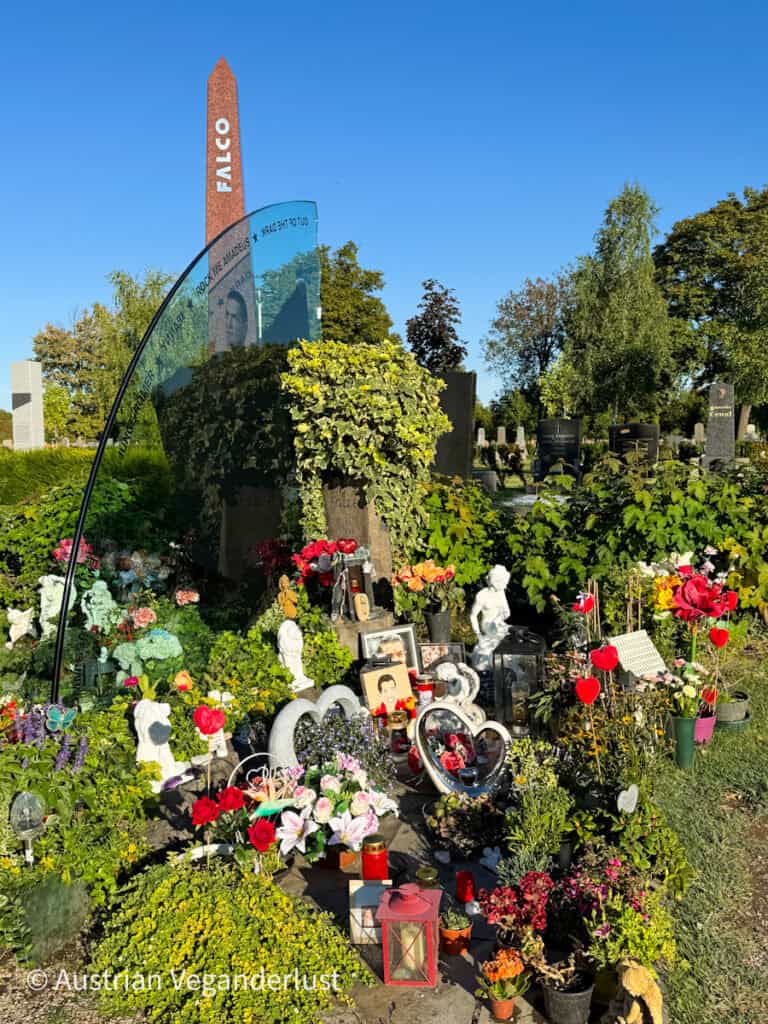 Farbenfrohes und reich geschmücktes Grab des österreichischen Musikers Falco mit Blumen, herzförmigen Ehrungen, Fotos und einem roten Obelisken, auf dem sein Name steht.