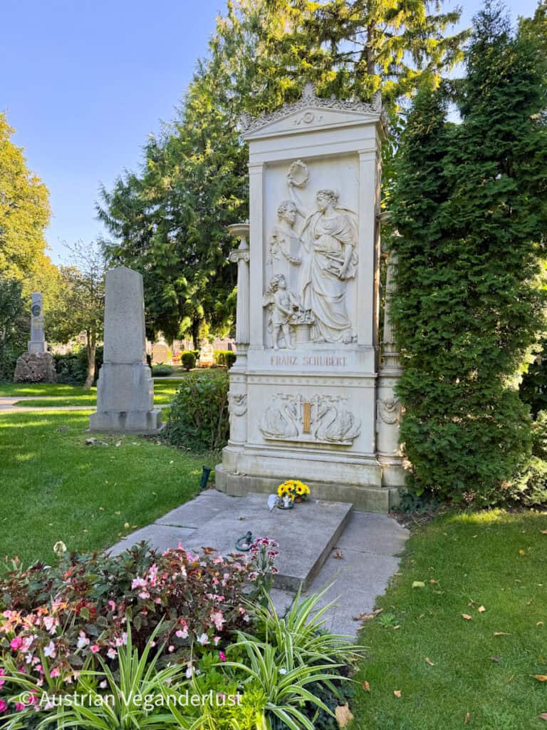 Der weiße Marmorgrabstein des Komponisten Franz Schubert ist mit Skulpturen verziert und von Grün und Blumen umgeben.