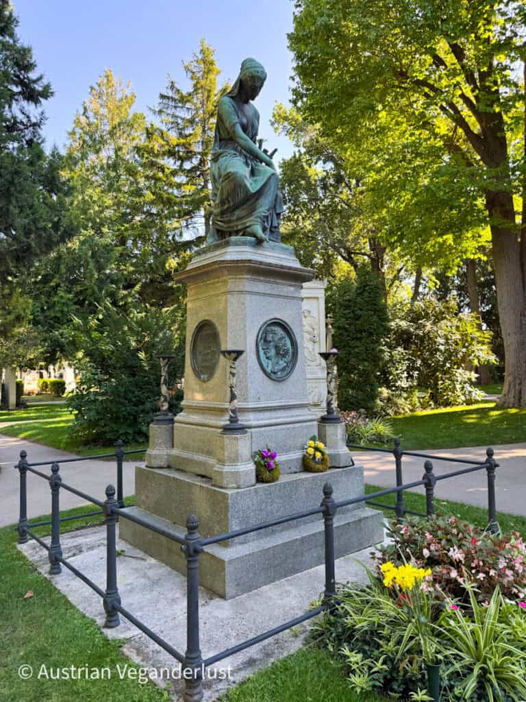 Monumentaler Grabstein mit einer Bronzestatue einer sitzenden Frau, umgeben von Blumenbeeten und Grünpflanzen auf dem Wiener Zentralfriedhof.
