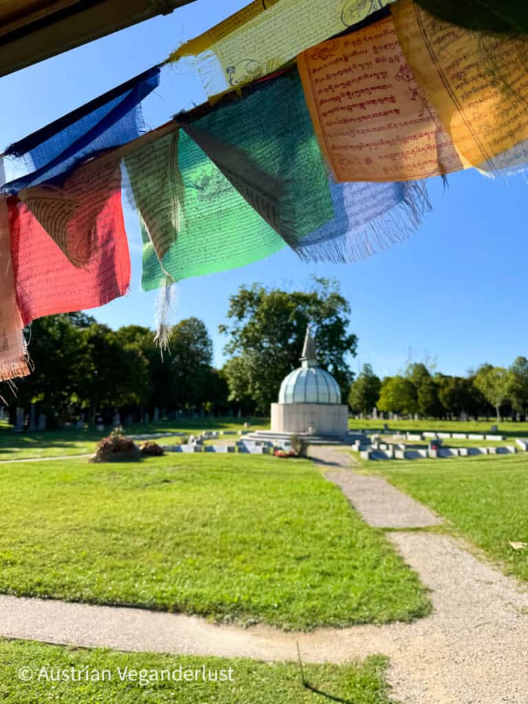 Eine Reihe farbenfroher tibetischer Gebetsfahnen flattert im Vordergrund, während im Hintergrund eine buddhistische Gedenkstätte mit Kuppel auf dem Wiener Zentralfriedhof zu sehen ist.