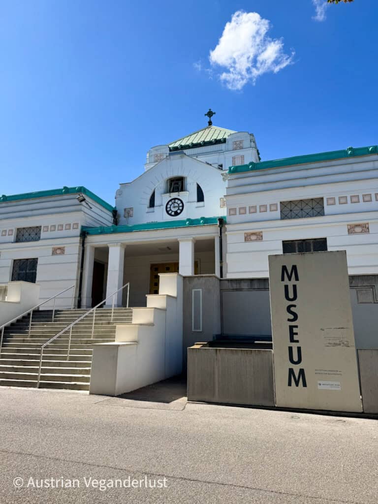 Außenansicht des Bestattungsmuseums am Wiener Zentralfriedhof mit weißer Fassade, grünem Dach, zentraler Uhr und einem großen Schild mit der Aufschrift "MUSEUM".
