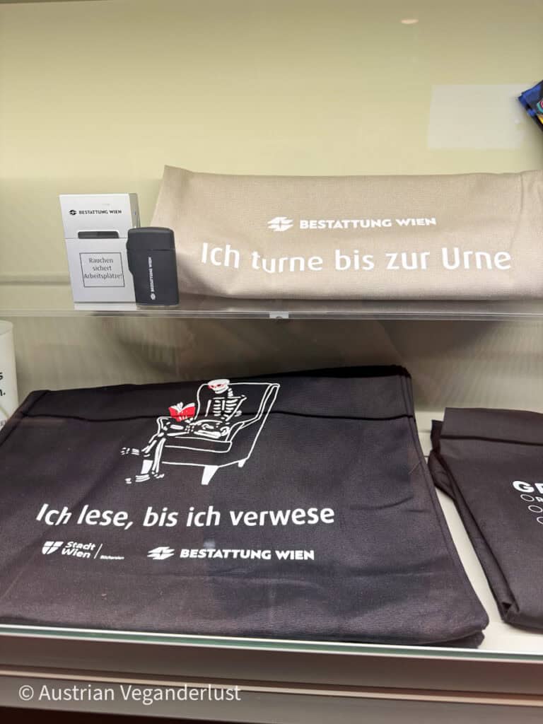 Ausstellungsregal im Bestattungsmuseum Wien mit Scherzartikeln von Bestattung Wien, darunter Tragetaschen mit witzigen Sprüchen wie "Ich turne bis zur Urne" und "Ich lese, bis ich verwese" sowie Markenfeuerzeuge und Zigarettenschachteln.