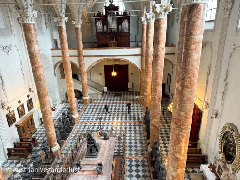 Eine erhöhte Ansicht der Hofkirche in Innsbruck zeigt schwarz-weiß karierte Böden, hohe rosa Marmorsäulen und ein zentrales Grabmal, das von lebensgroßen Bronzestatuen historischer Persönlichkeiten umgeben ist, mit einer Orgelempore über dem Eingang.