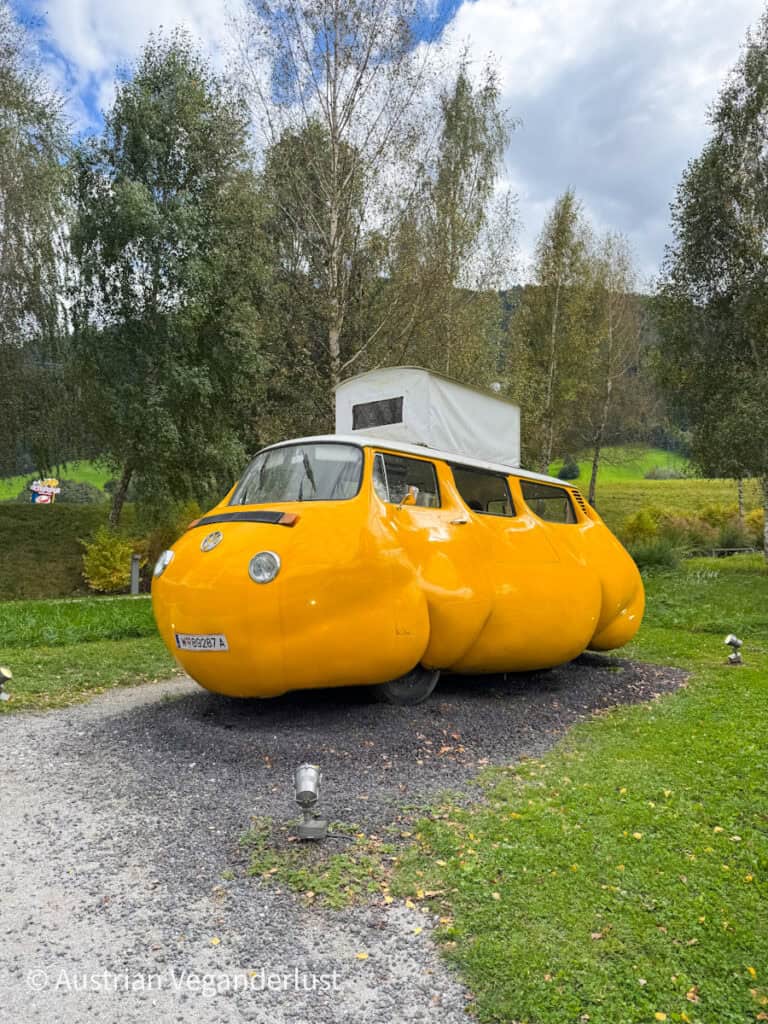 Playful sculpture of a chubby yellow VW-style camper van with exaggerated round curves, parked on gravel in a green park surrounded by birch trees.