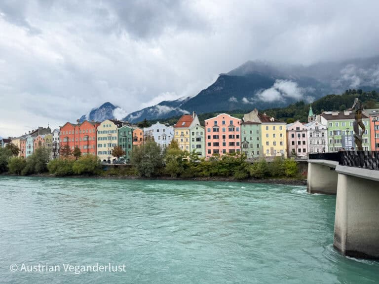 Row of pastel-colored buildings along the turquoise Inn River in Innsbruck, backed by foggy mountain peaks and overcast skies.