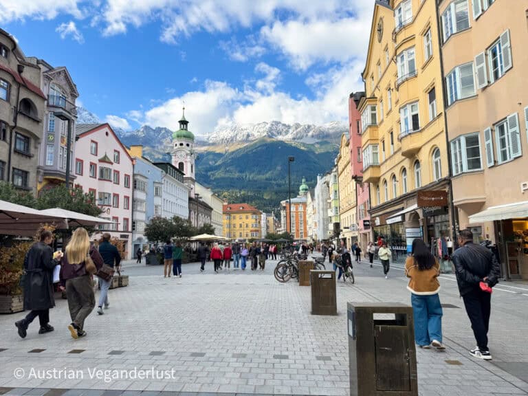 Bustling pedestrian street in Innsbruck lined with colorful historic buildings and shopfronts, with snow-capped mountains rising in the background under a blue sky.