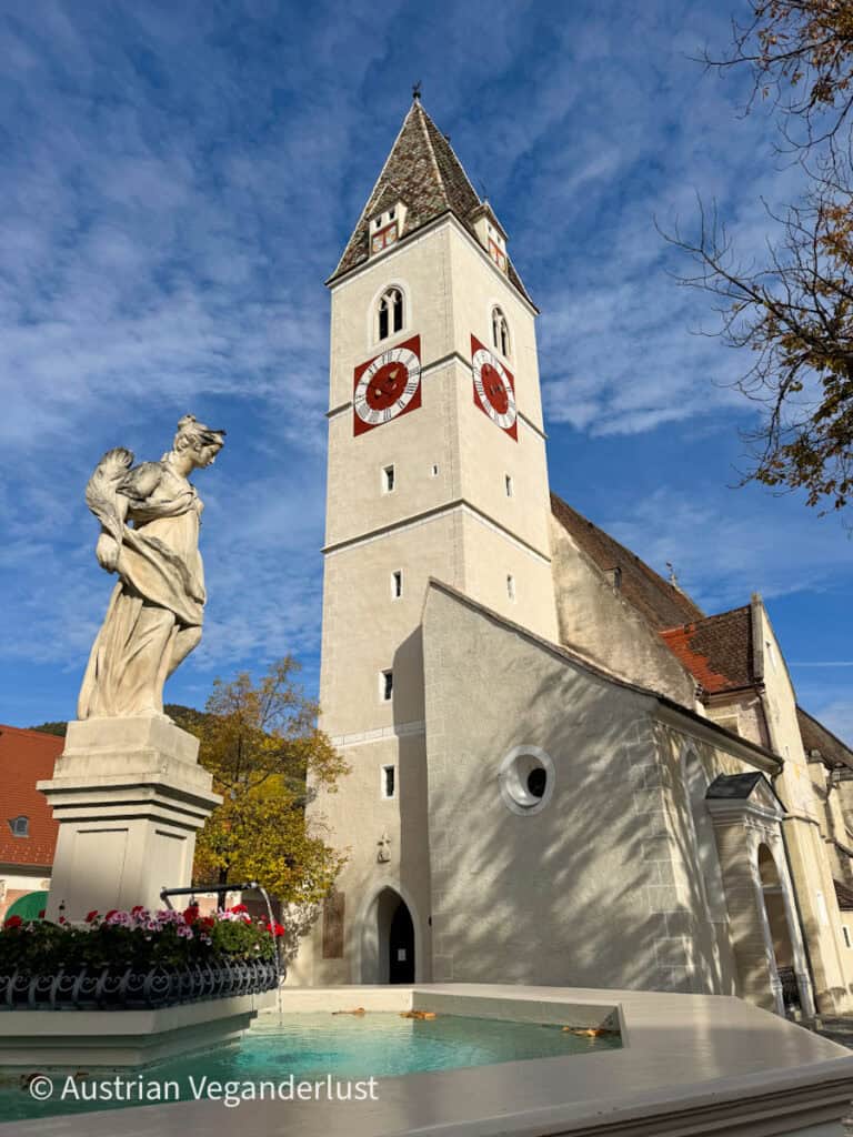A tall white church tower with red and white clock faces stands against a blue sky, beside a stone statue and small fountain decorated with flowers.