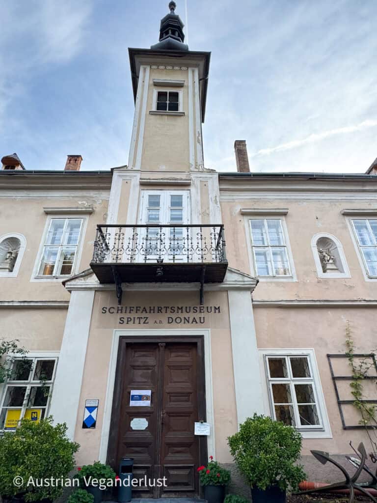 Front entrance of the Schifffahrtsmuseum Spitz a.d. Donau, a beige historic building with a central clock tower and decorative balcony above the wooden door.