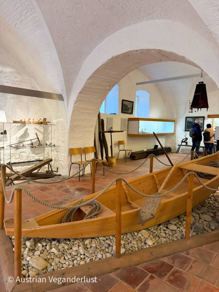 An arched stone room inside a museum featuring a small wooden boat display surrounded by ropes and pebbles, with historical tools and artifacts on the walls.