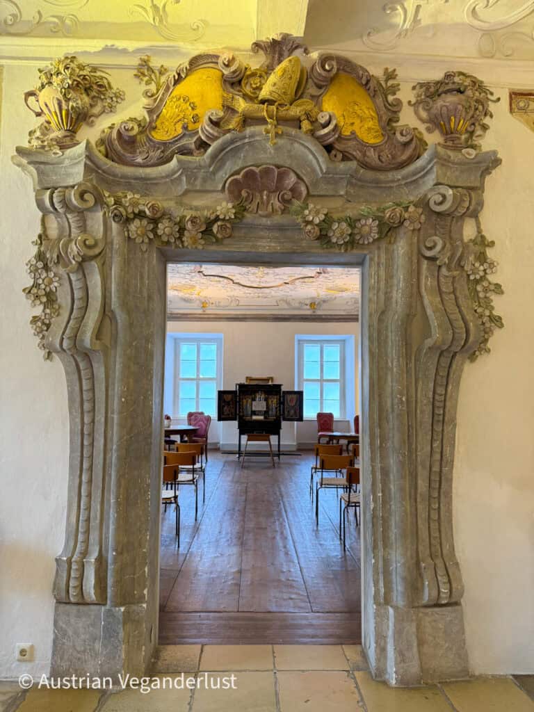 A lavishly carved baroque-style doorway adorned with golden religious symbols and floral motifs opens into a museum room with wooden floors and antique furniture.