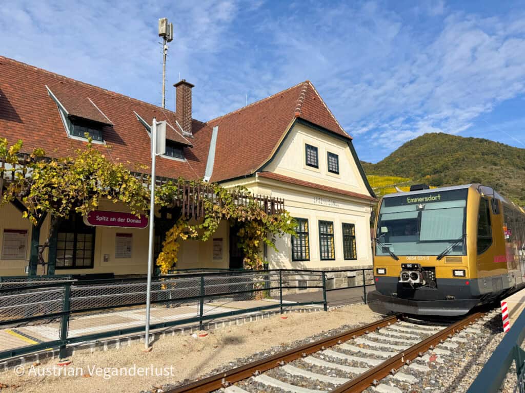 A gold-colored train bound for Emmersdorf is parked at the charming Spitz an der Donau train station, surrounded by hills and grapevines.