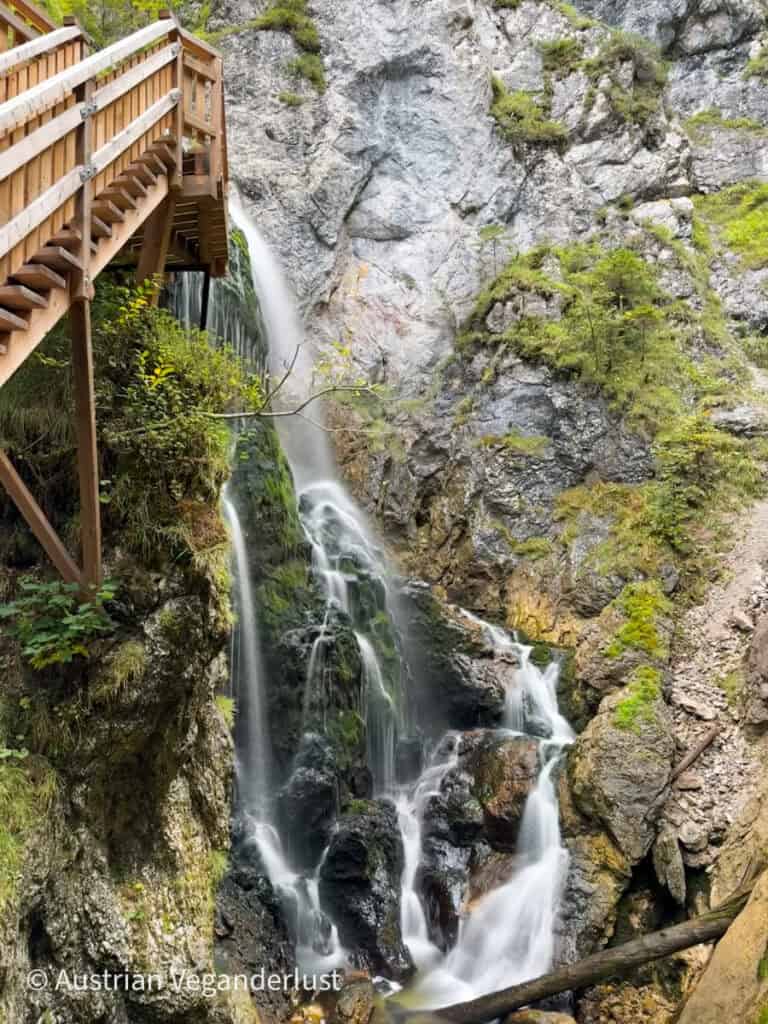Ein anmutiger Wasserfall ergießt sich neben einer hölzernen Aussichtsplattform, die über der Klippe schwebt, über moosbewachsene Felsschichten, eingerahmt von schroffem Berggelände.