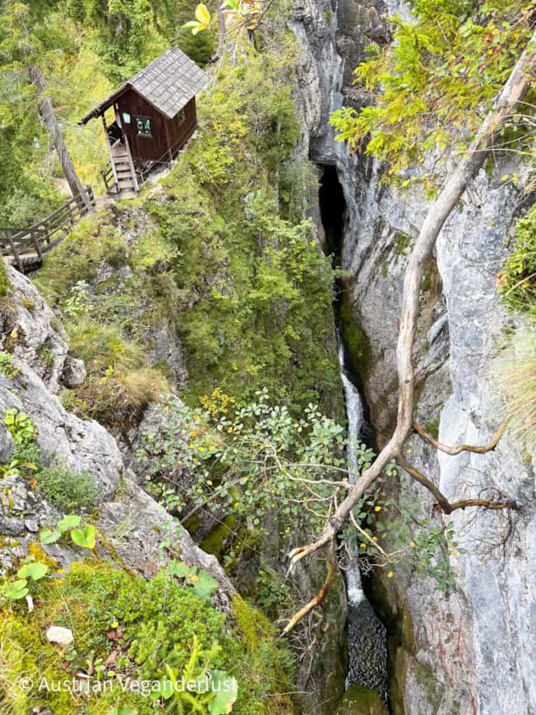 Eine kleine Holzhütte steht bedenklich nahe am Rand einer tiefen Felsspalte, wo ein schmaler Wasserfall in der Dunkelheit verschwindet.