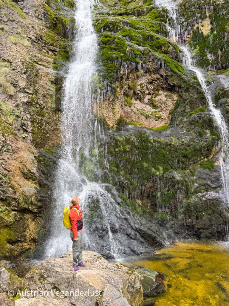 Eine Frau mit einem gelben Rucksack steht auf einem Felsen und bewundert einen hohen Wasserfall, der über moosbewachsene Klippen in ein seichtes, goldenes Becken stürzt.