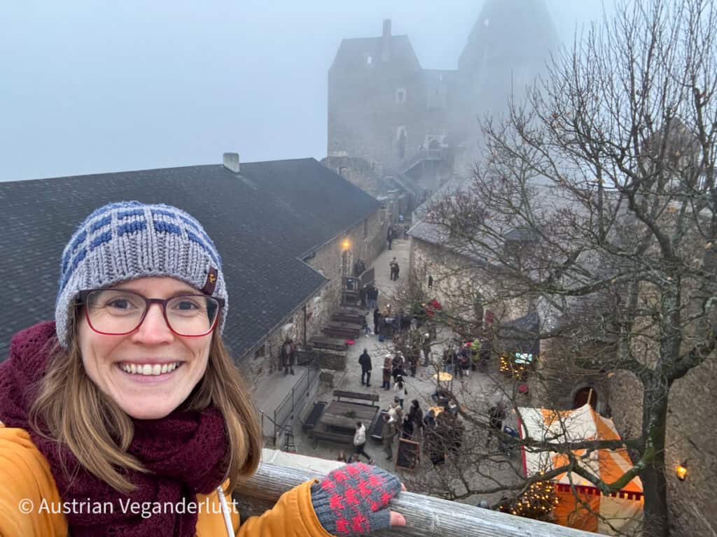Tina in a knit hat and scarf takes a selfie on a foggy day overlooking the medieval courtyard of Aggstein Castle filled with Christmas market stalls.
