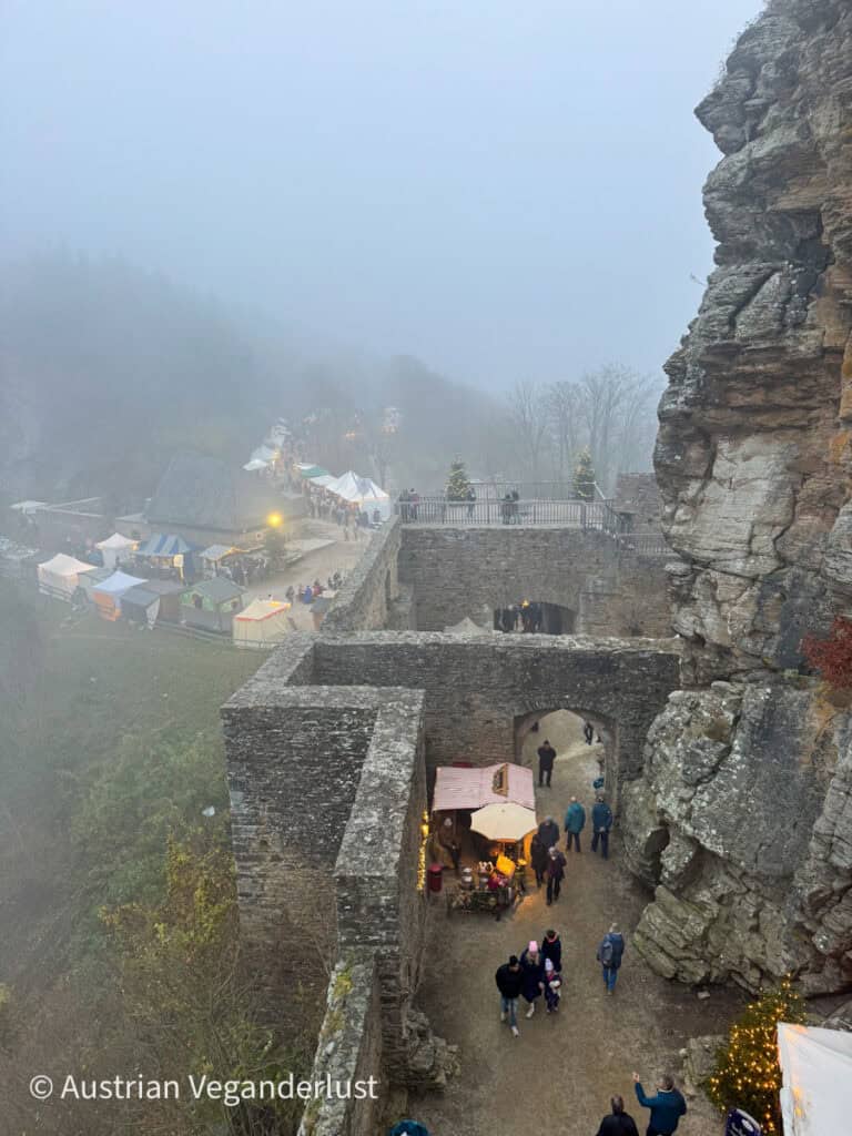 Foggy view of a Christmas market behind stone castle walls with tents, Christmas trees, and visitors exploring the medieval setting
