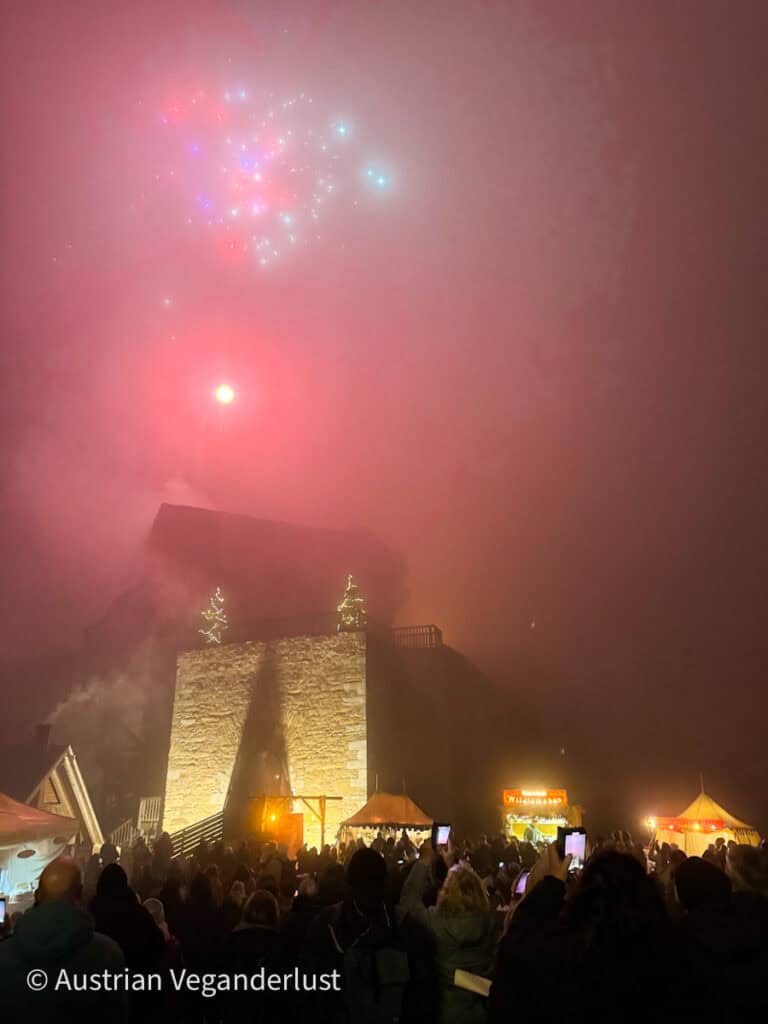 Nighttime fireworks above a foggy castle entrance at Burg Aggstein with a crowd watching and Christmas lights glowing