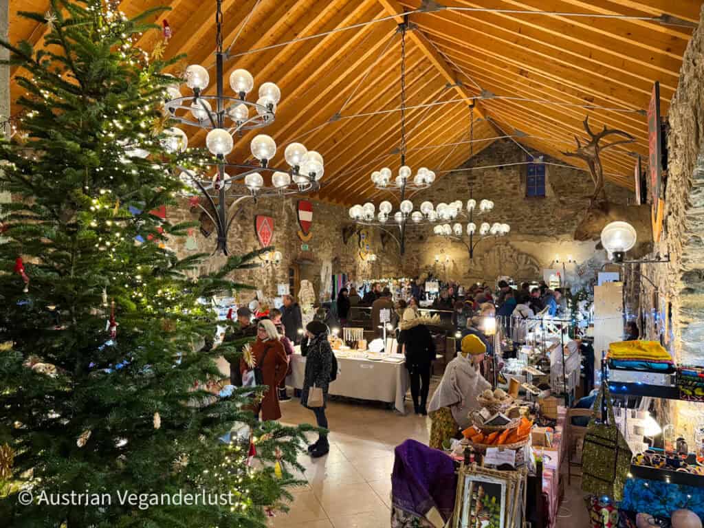 Festive indoor market inside a stone hall with wooden beams, chandeliers, decorated stalls, and a large Christmas tree